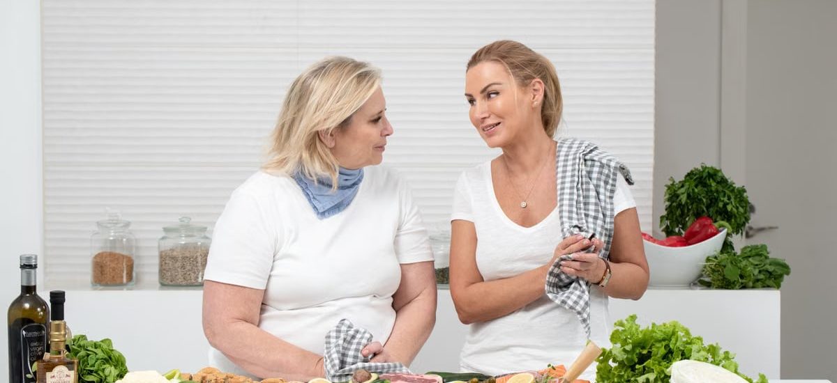 Two women in a kitchen preparing fresh vegetables and cheese with olive oil and spices.