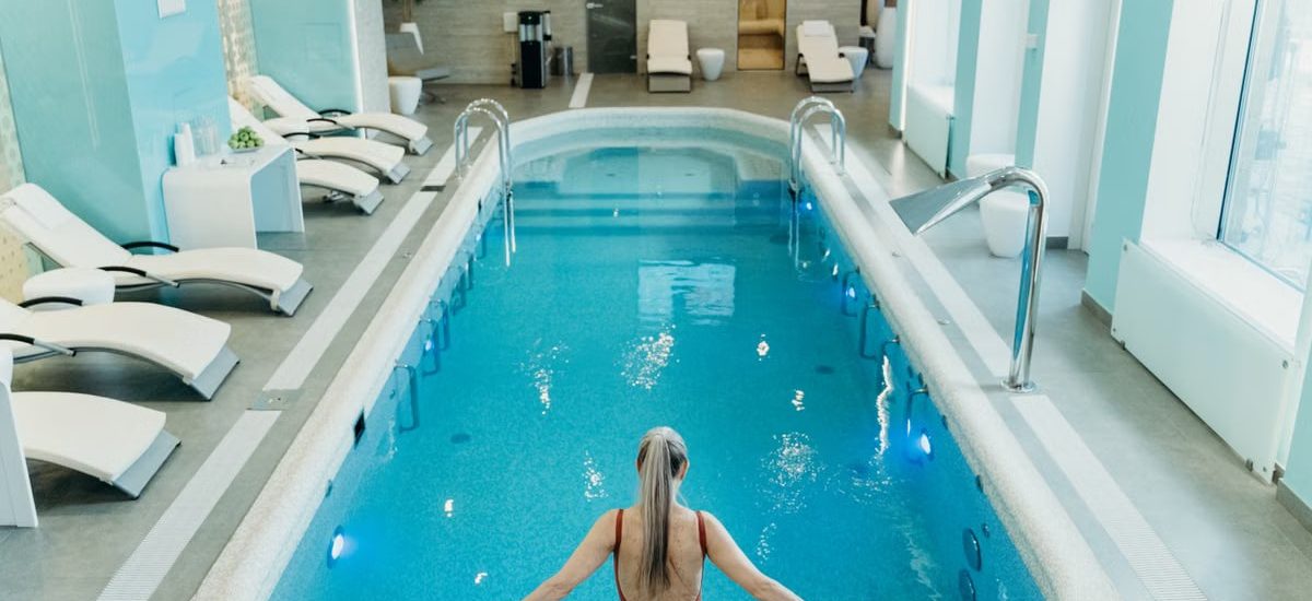 A woman in a red swimsuit prepares to enter an elegant indoor pool at a luxurious spa.